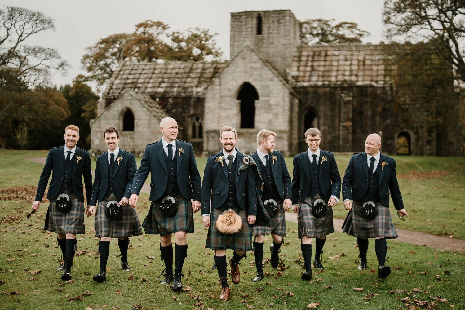 Men in formal kilts and suits pose together on grass with stone buildings in background.
