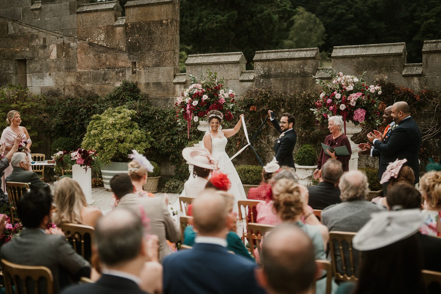 Wedding guests seated in wooden chairs during an outdoor ceremony with stone architecture.