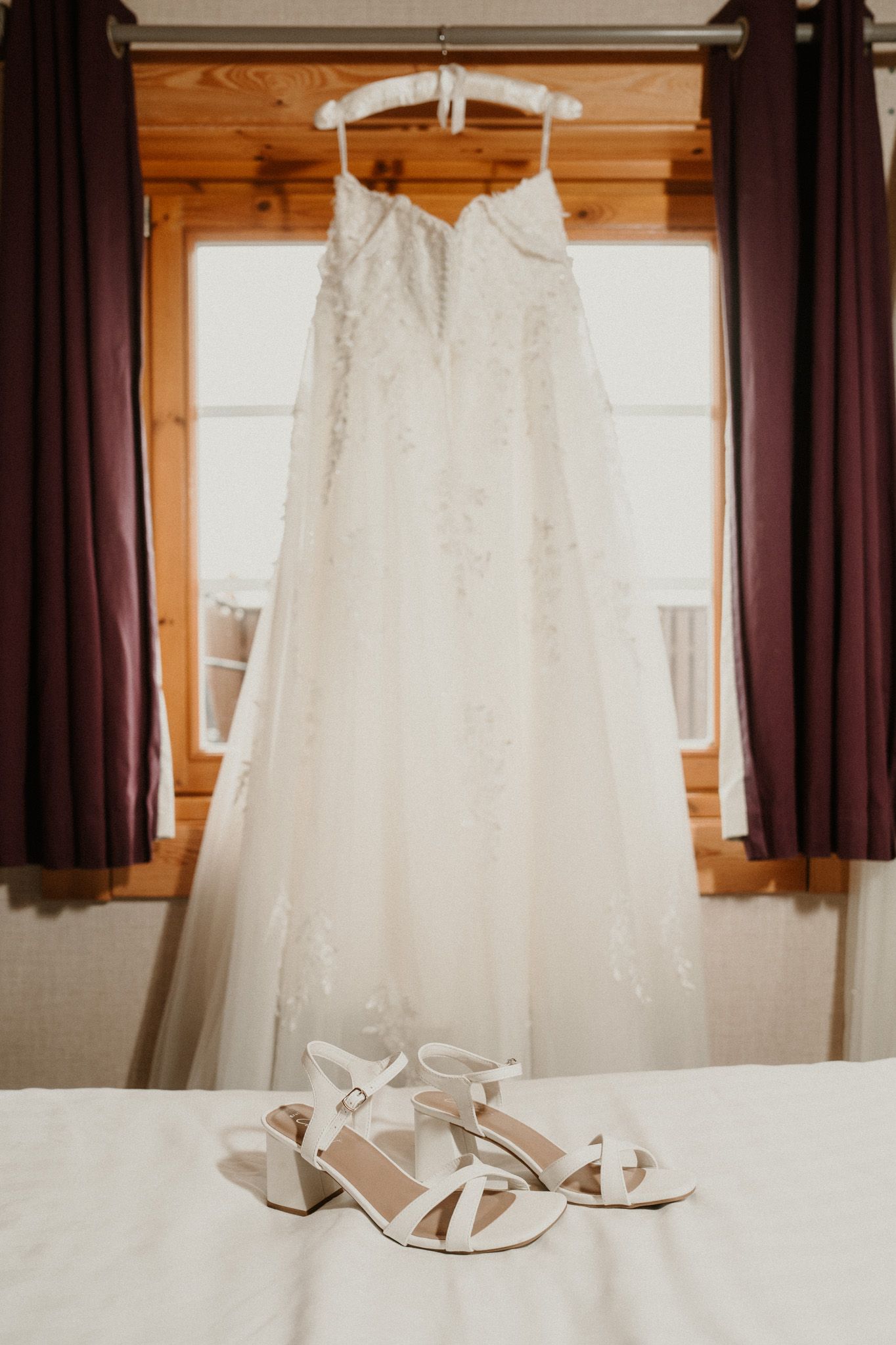 White wedding dress and shoes displayed hanging against wooden walls and burgundy curtains.