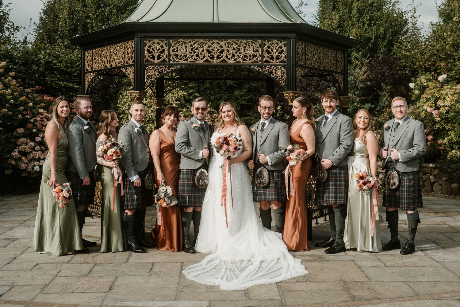 Full wedding party portrait with groomsmen and bridesmaids under outdoor pavilion.