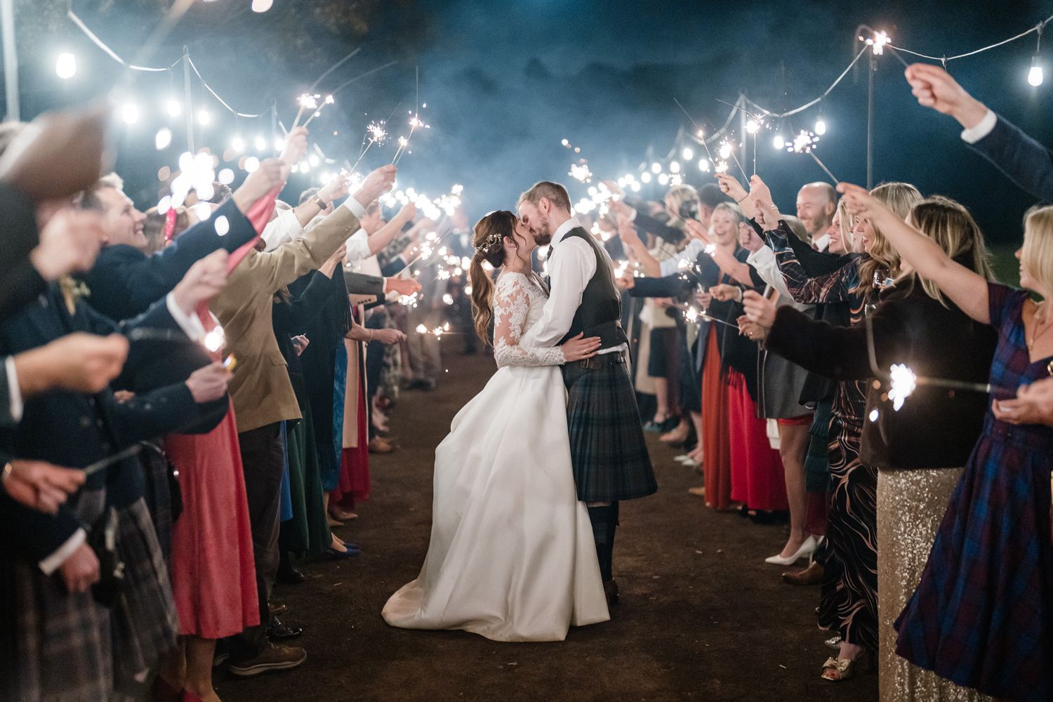 Sparklers create a magical glow as wedding guests celebrate under twinkling string lights.