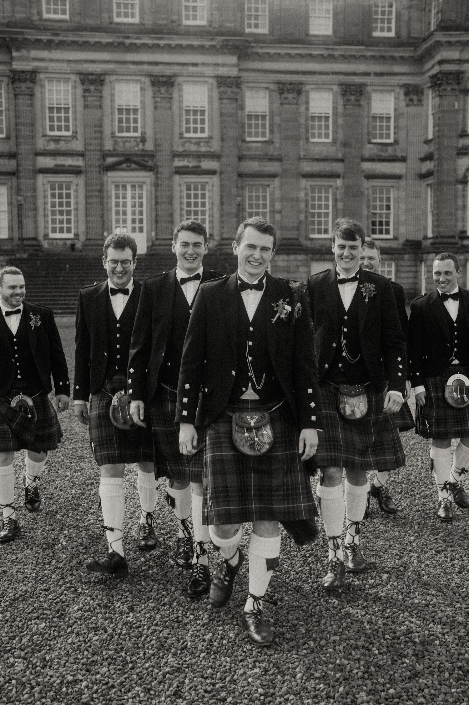 Groomsmen in traditional Scottish attire with kilts stand in a line outside a historic stone building.