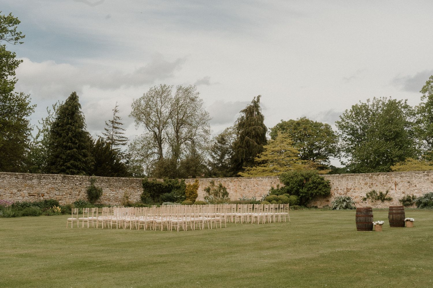 Outdoor wedding ceremony setup with white chairs on green lawn, stone wall backdrop, and wooden barrels under cloudy sky.