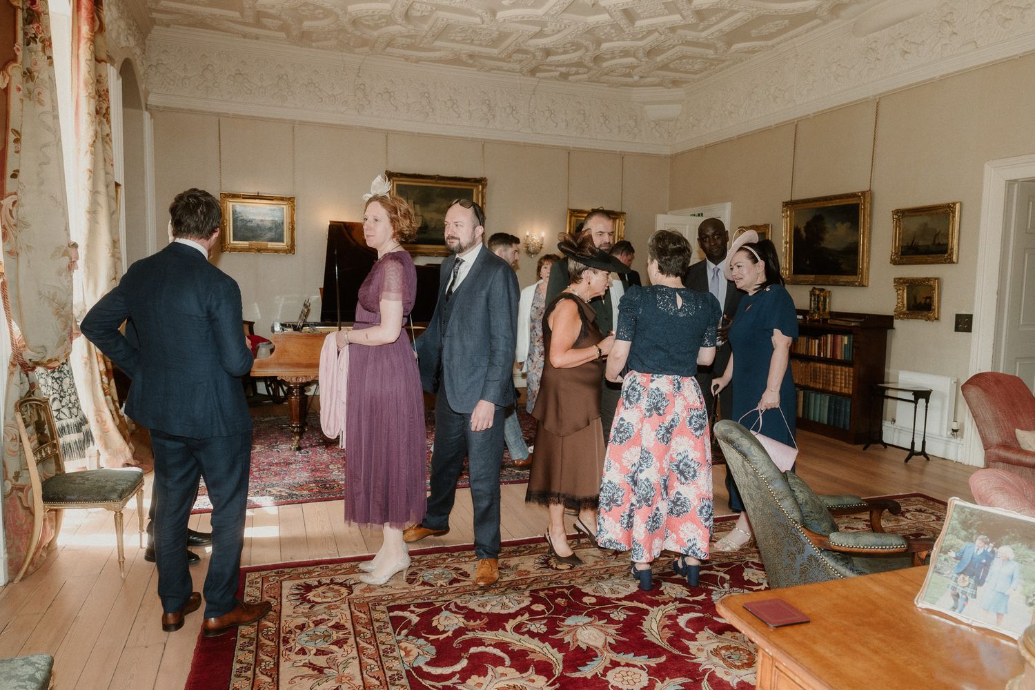 Guests mingling in an elegant room with ornate decor, red rug, and paintings during a formal gathering.
