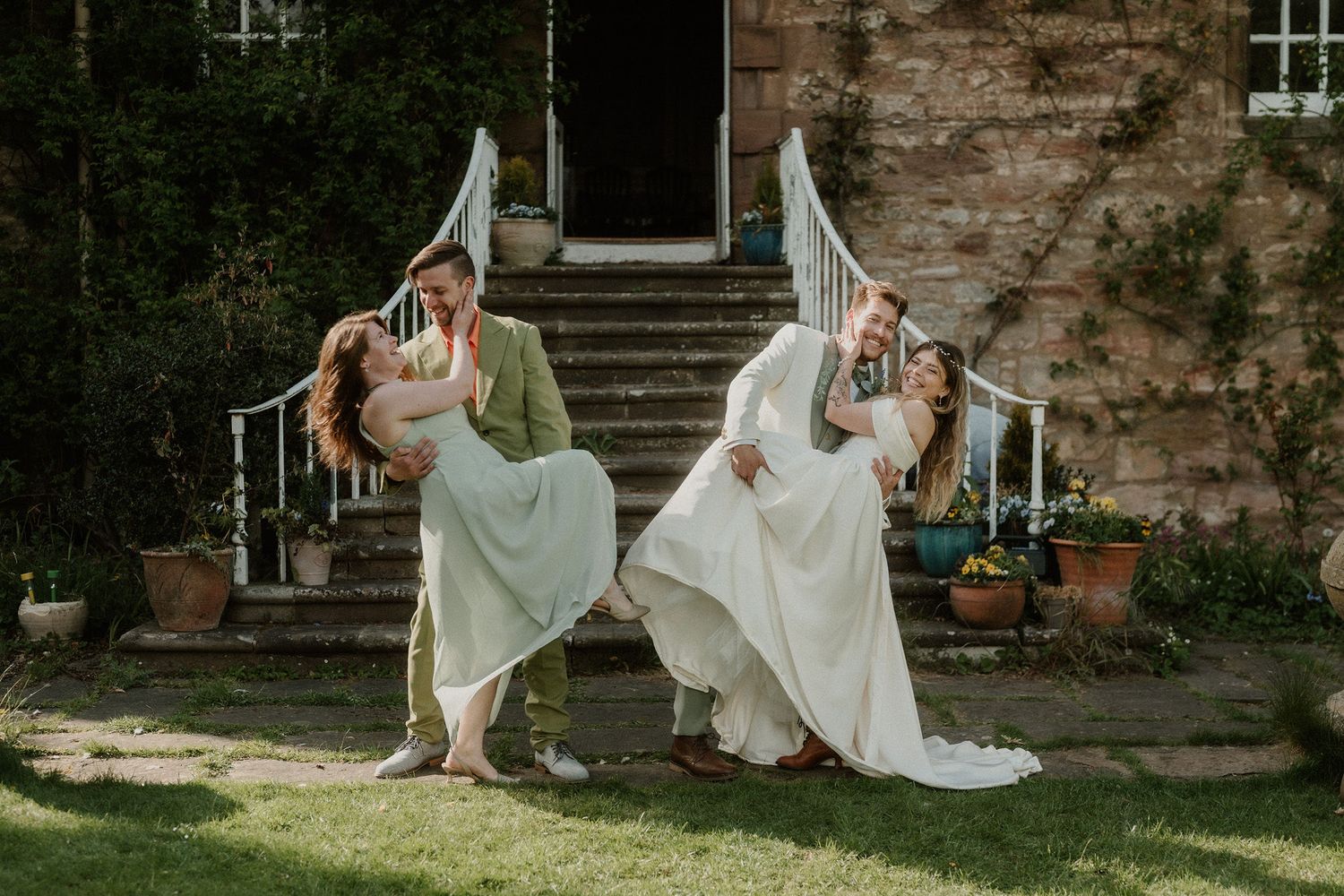 Two couples share romantic dips in front of a charming stone manor, dressed in wedding attire on a sunny day.