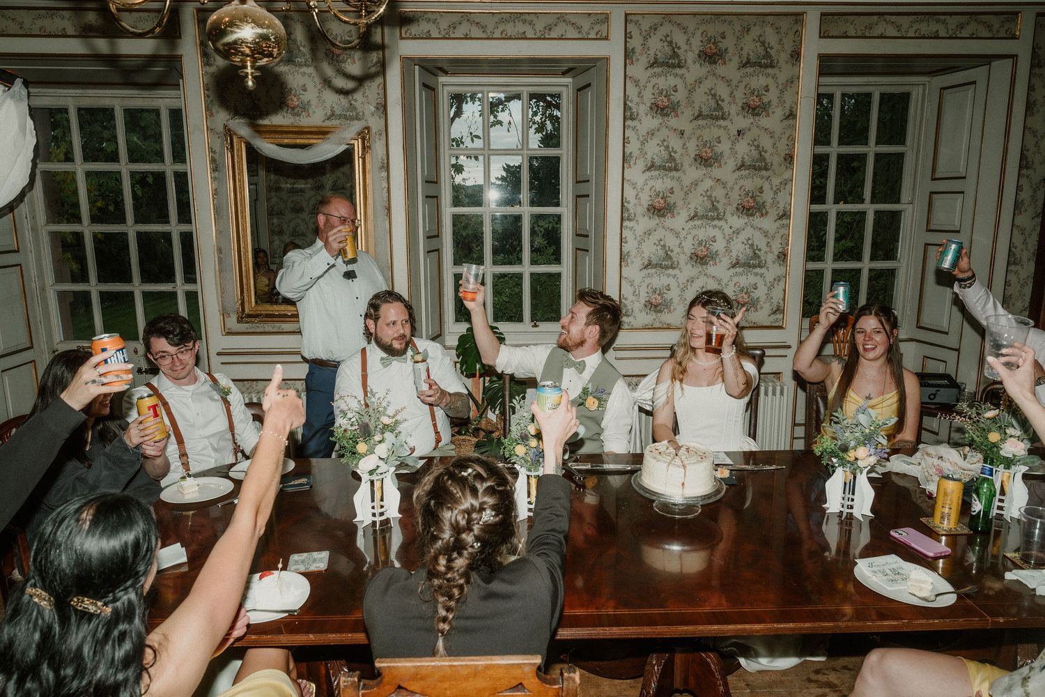 Wedding reception toast with bride and groom at elegant dining table surrounded by cheering guests in decorated room.