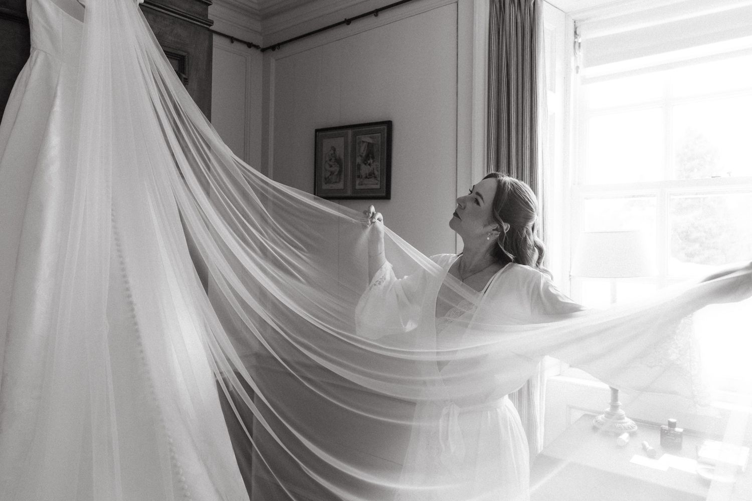 Black and white photo of a bride holding up her flowing wedding veil near a bright window before her ceremony.
