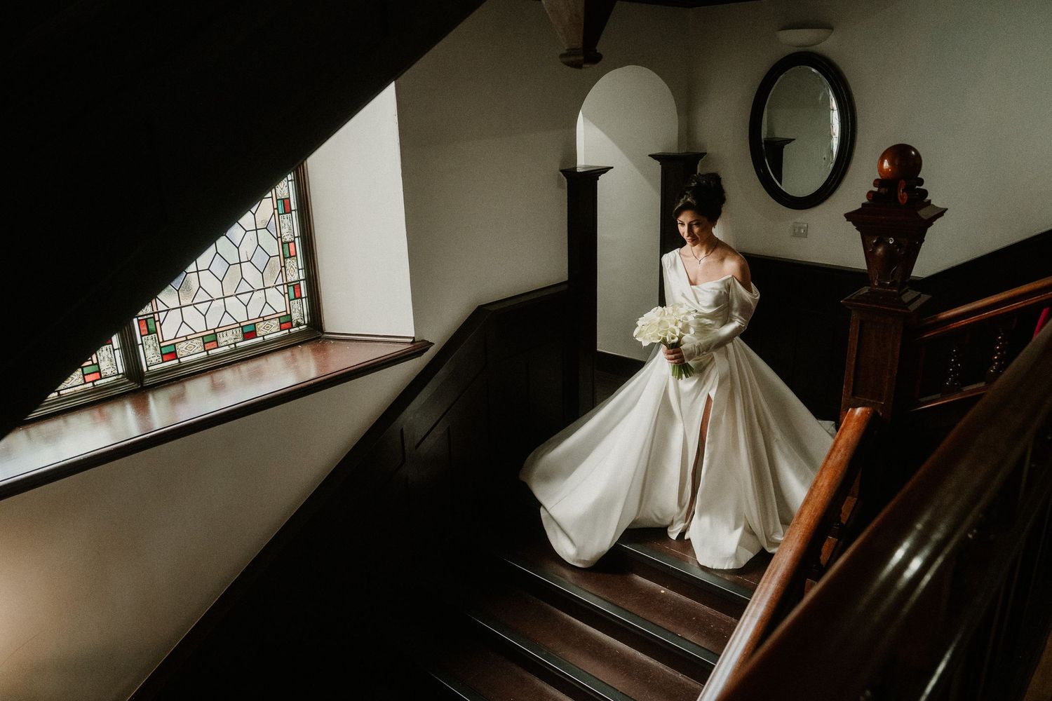 A bride in a white wedding gown descends an elegant dark wooden staircase in dramatic lighting.