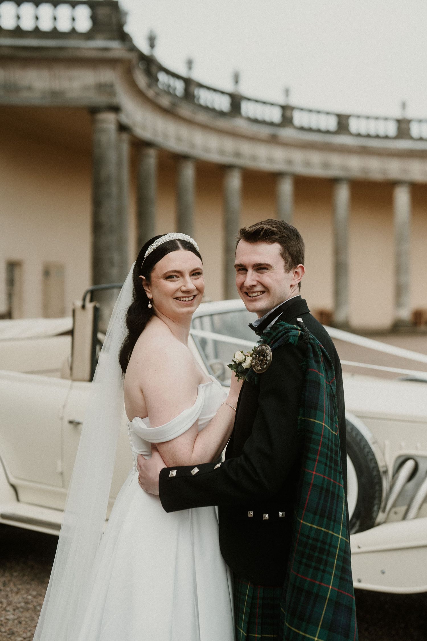 Wedding couple shares intimate moment near vintage car at Scottish estate.