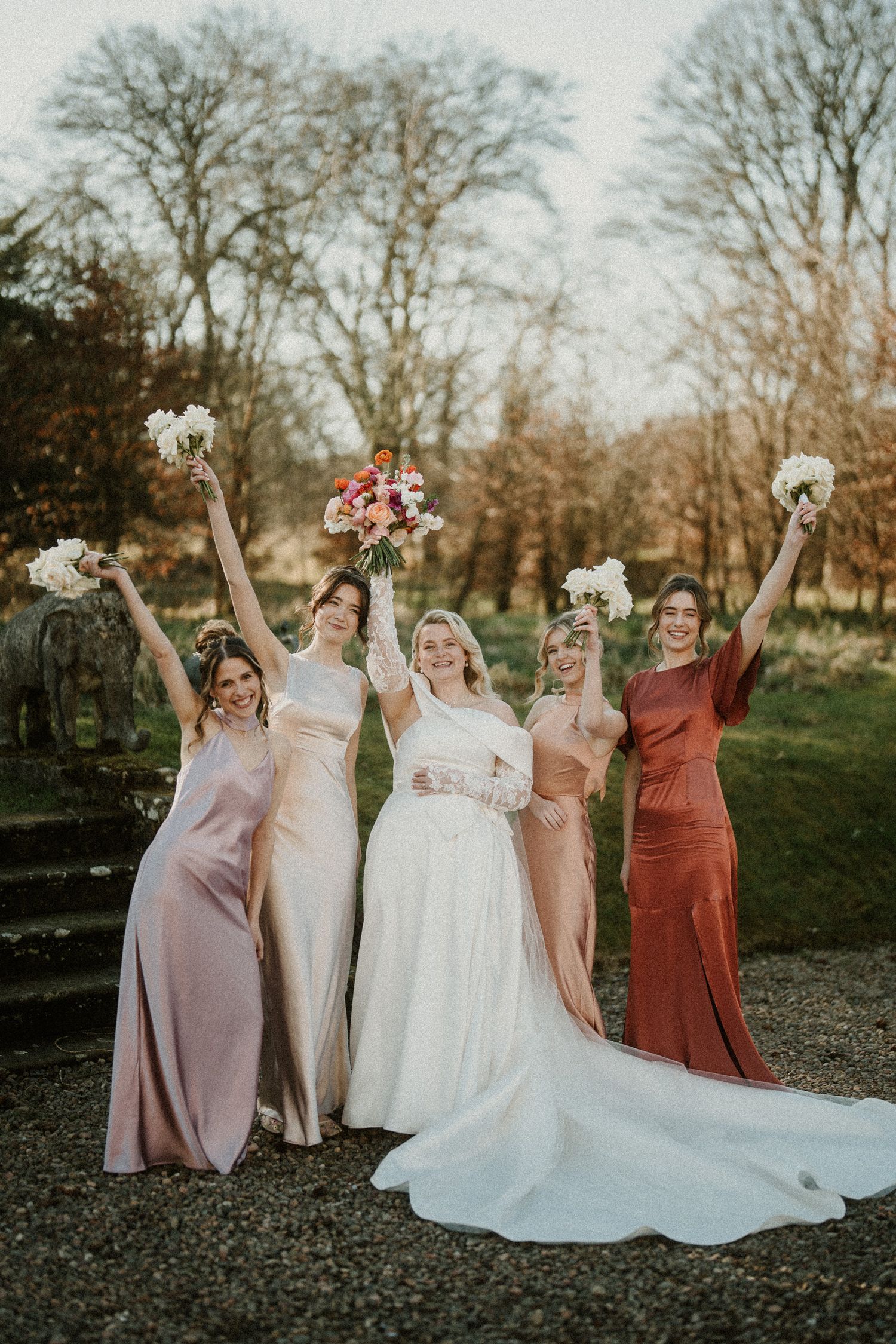 Wedding party celebrates joyfully with raised arms in coordinated pink and rust dresses against autumn backdrop.