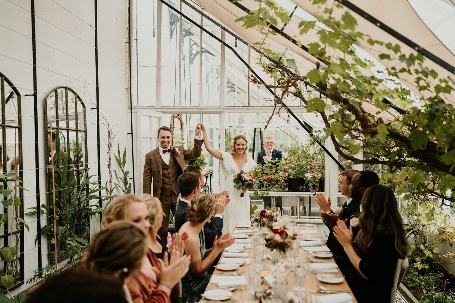 Wedding celebration inside an elegant greenhouse with guests seated at long tables.