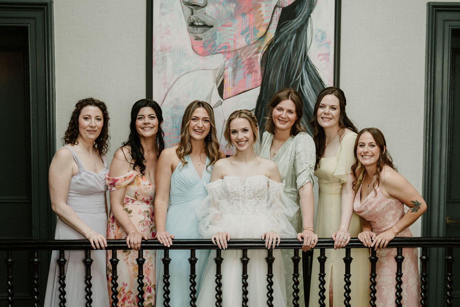 Bridesmaids in soft pastel dresses lined up along ornate balcony railing.