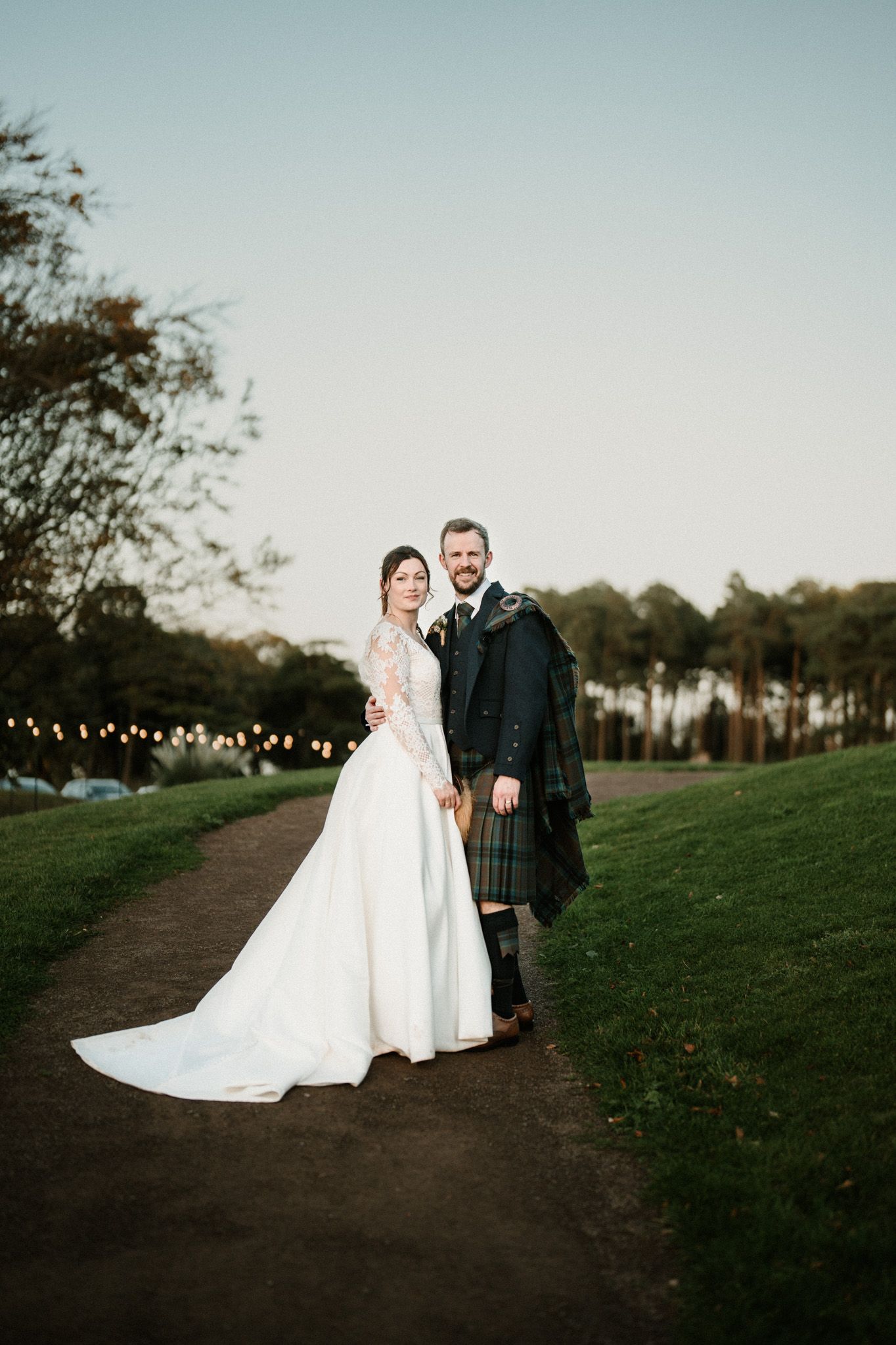 Couple in wedding attire walking along a scenic outdoor path at sunset.