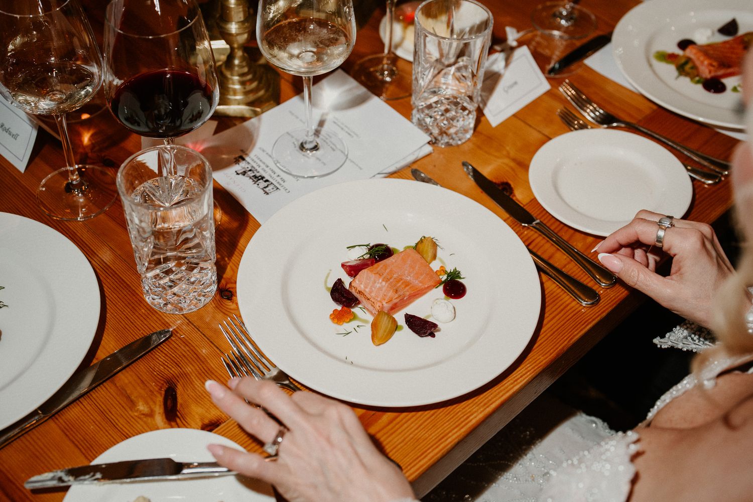 Wedding guests enjoy an elegant dinner at a wooden table set with white plates and crystal glasses.