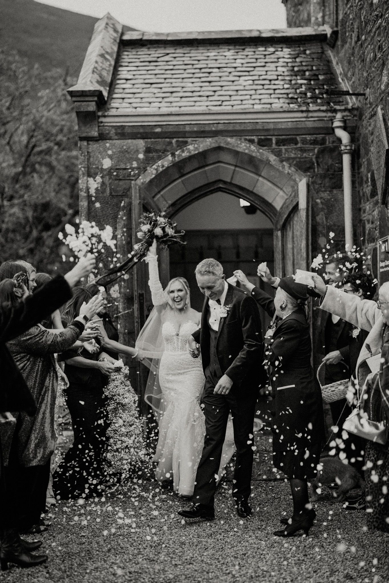 Black and white photo of newlyweds leaving church under shower of confetti.
