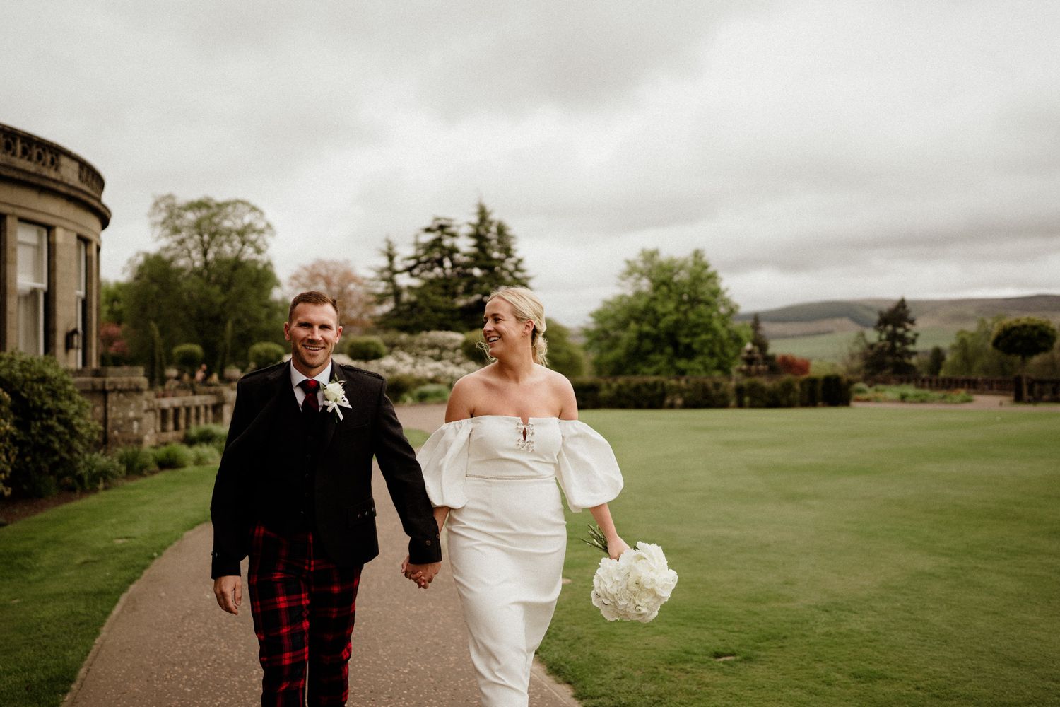 Elegant wedding couple poses together on a sprawling green lawn outside a historic stone building.