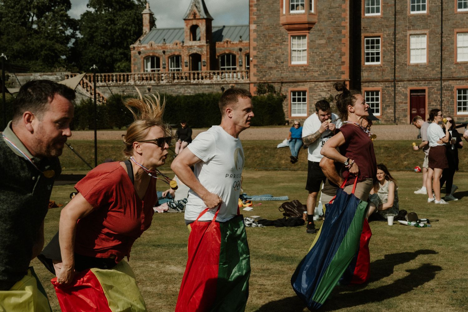 Adults in colorful sacks compete in a sack race on a sunny lawn outside a brick building with onlookers watching.