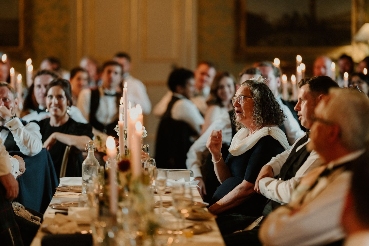 Guests seated at a long candlelit banquet table laugh and chat during an elegant formal dinner in a grand, ornate venue.