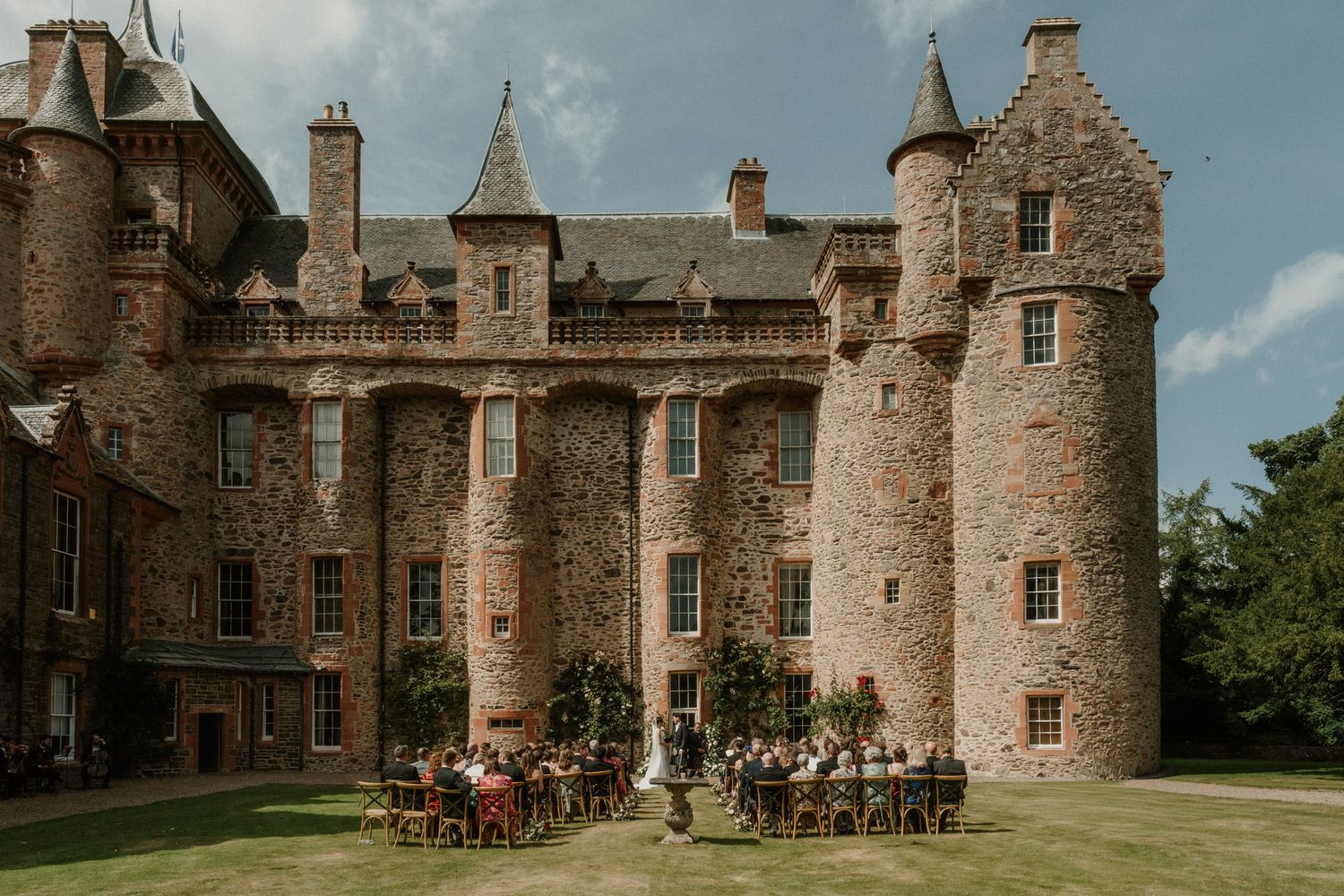 Outdoor wedding reception with guests seated at tables on a green lawn in front of a historic pink sandstone Scottish castle with turrets.