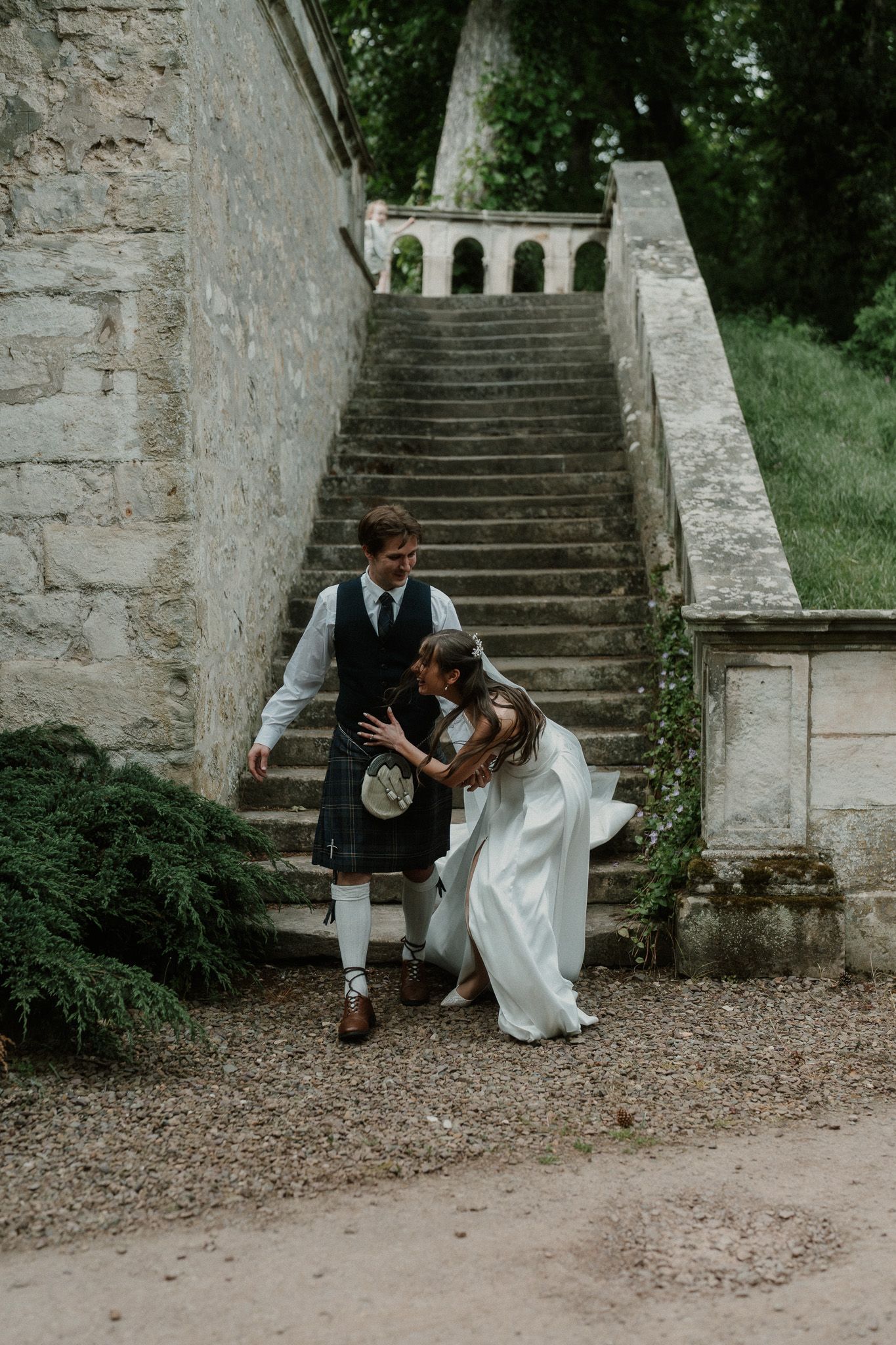 Bride and groom embrace near stone staircase, groom wearing traditional Scottish kilt at outdoor wedding venue.