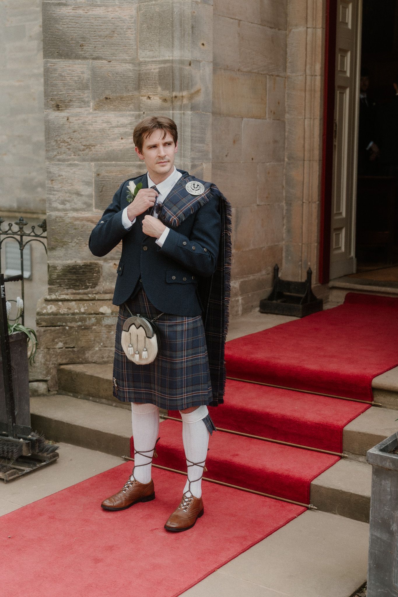 Man in traditional Scottish kilt outfit with sporran and ghillie brogues standing on red carpet steps.