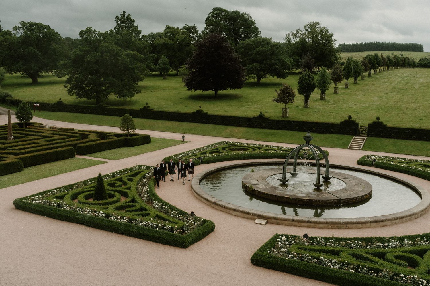 Aerial view of elegant formal garden with circular fountain, manicured hedges, and lush green landscape.