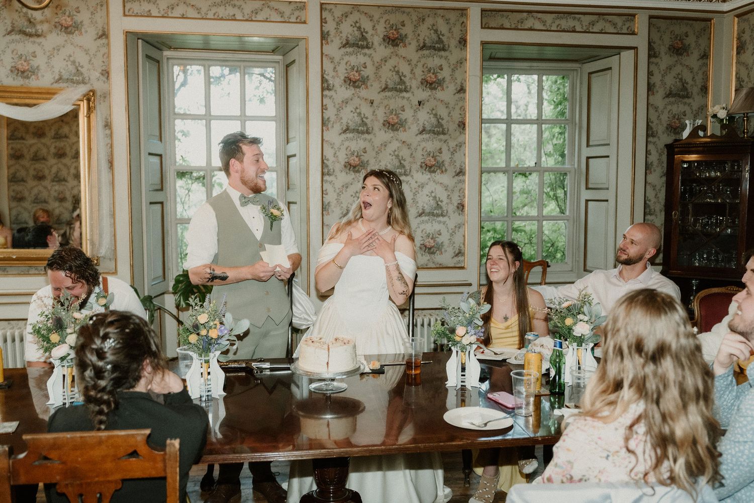 Bride and groom laughing joyfully during wedding reception dinner in elegant floral wallpapered dining room with guests.