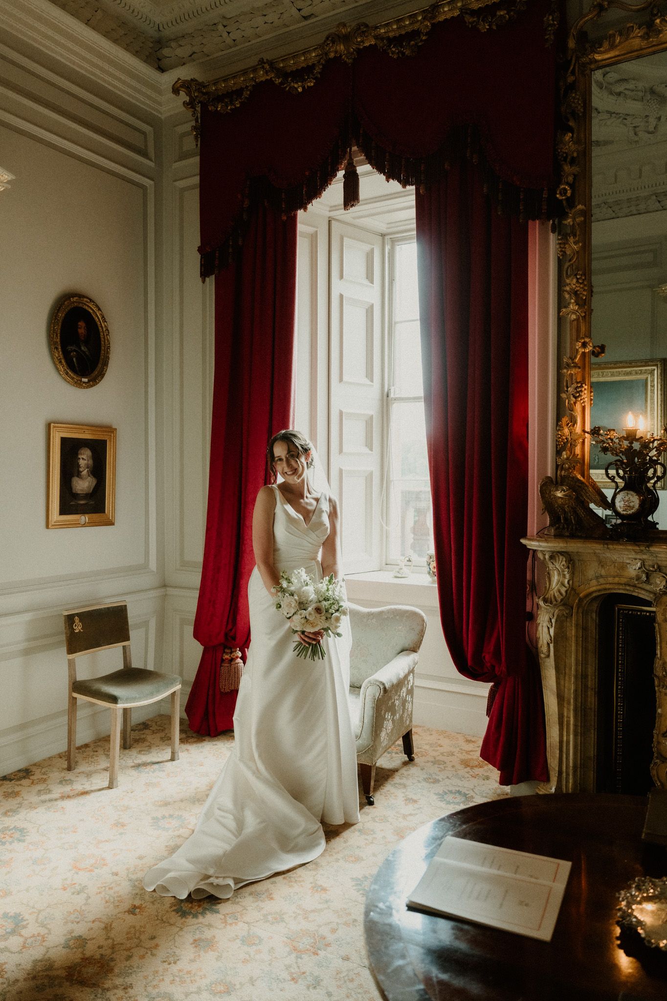 Bride in an elegant white gown holding a bouquet by a tall window with red velvet curtains in a grand room with a marble fireplace.