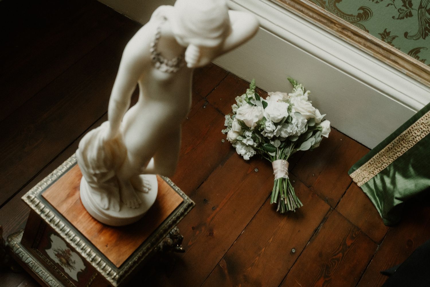 Overhead view of a white bridal bouquet with greenery on a wooden floor beside a marble statue on an ornate pedestal.