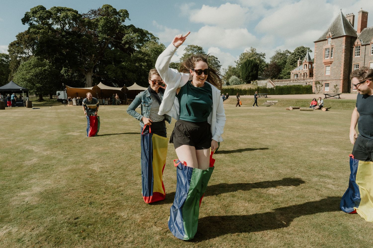 Women in colorful sacks hop across a sunny lawn during a fun sack race at an outdoor event near a historic stone manor.
