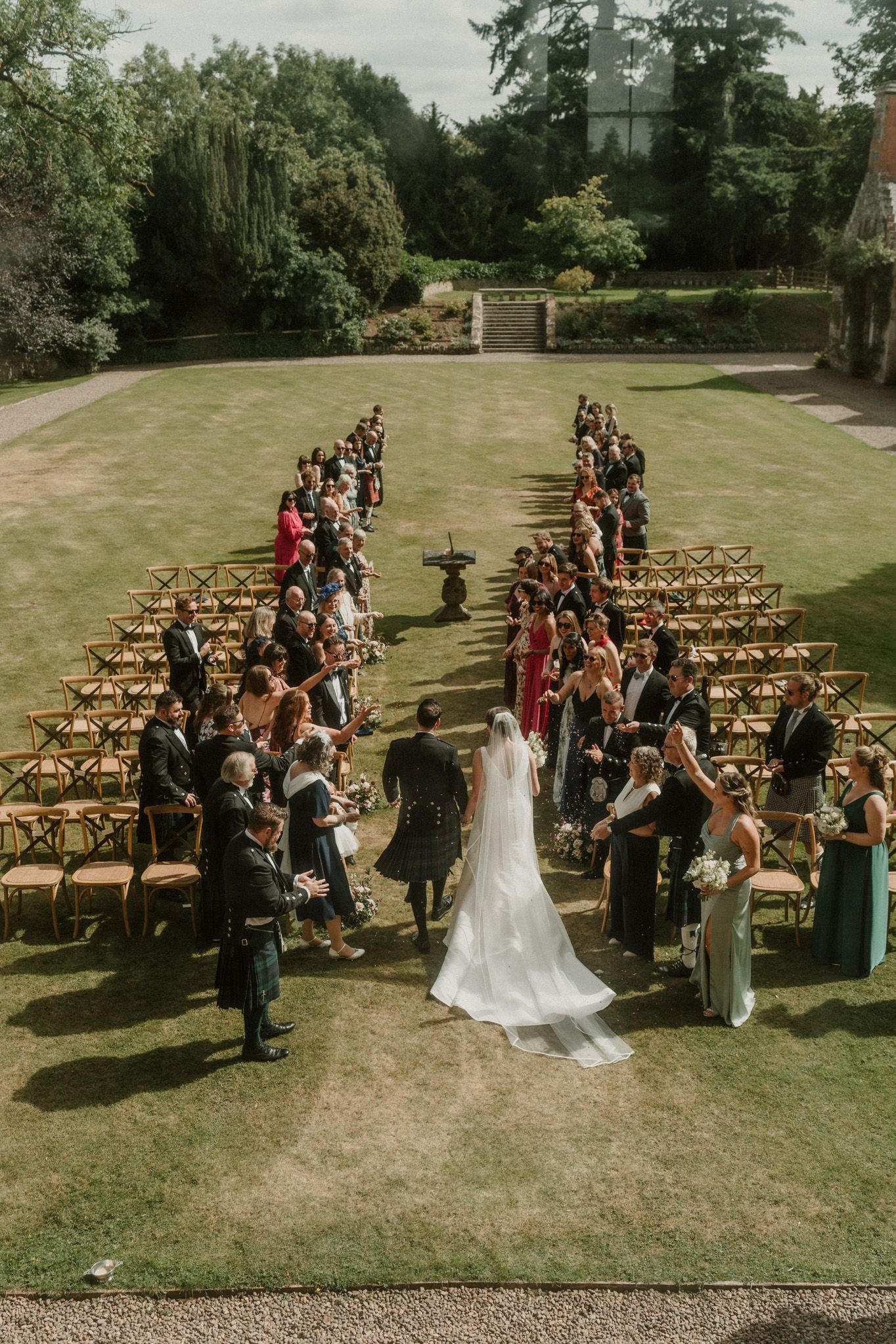 Bride in a long white gown and groom walk down the aisle between rows of guests at an outdoor garden wedding ceremony on a sunny lawn.