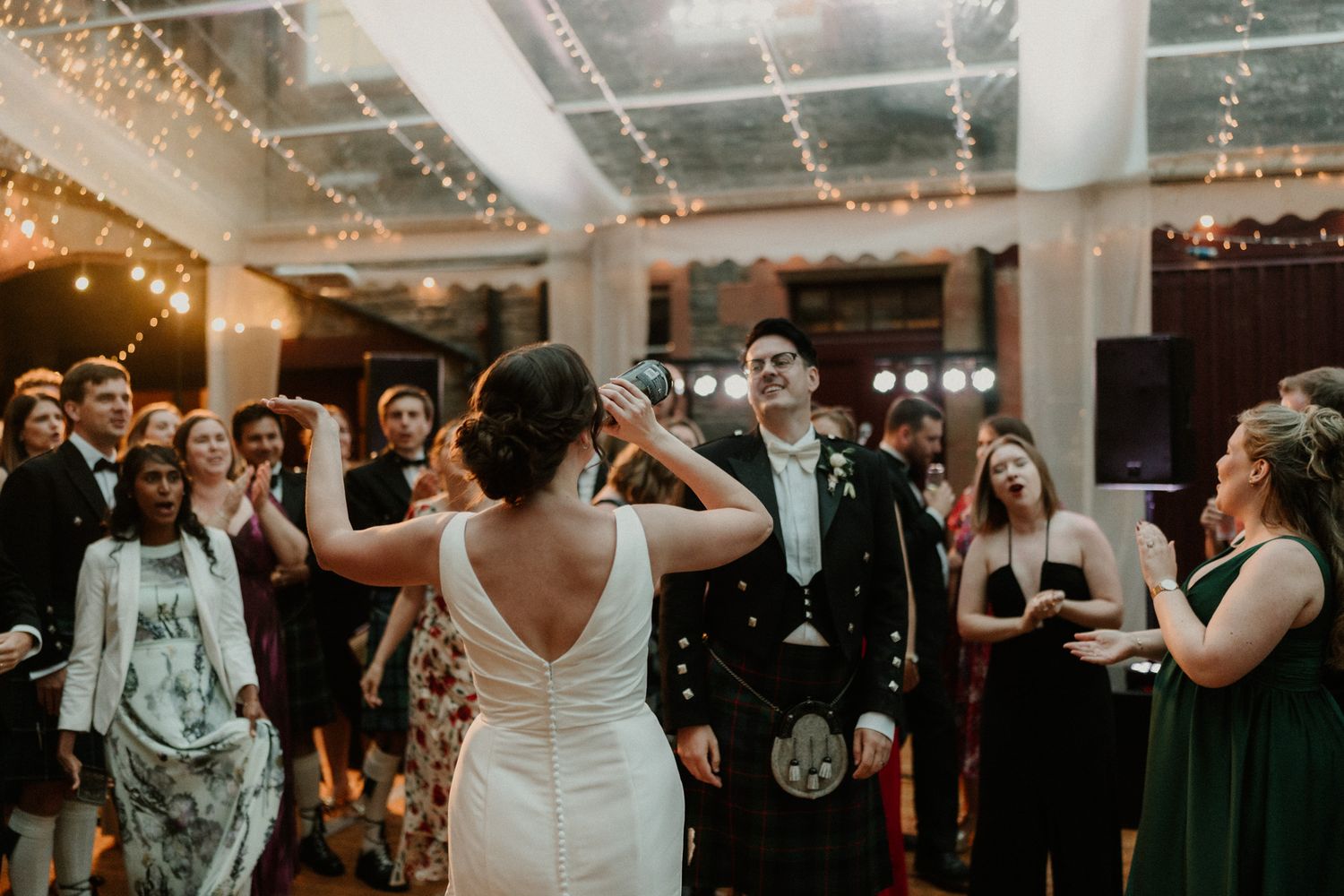 Bride in a white gown dances with arms raised toward a groom in a kilt as wedding guests cheer under a canopy of string lights.
