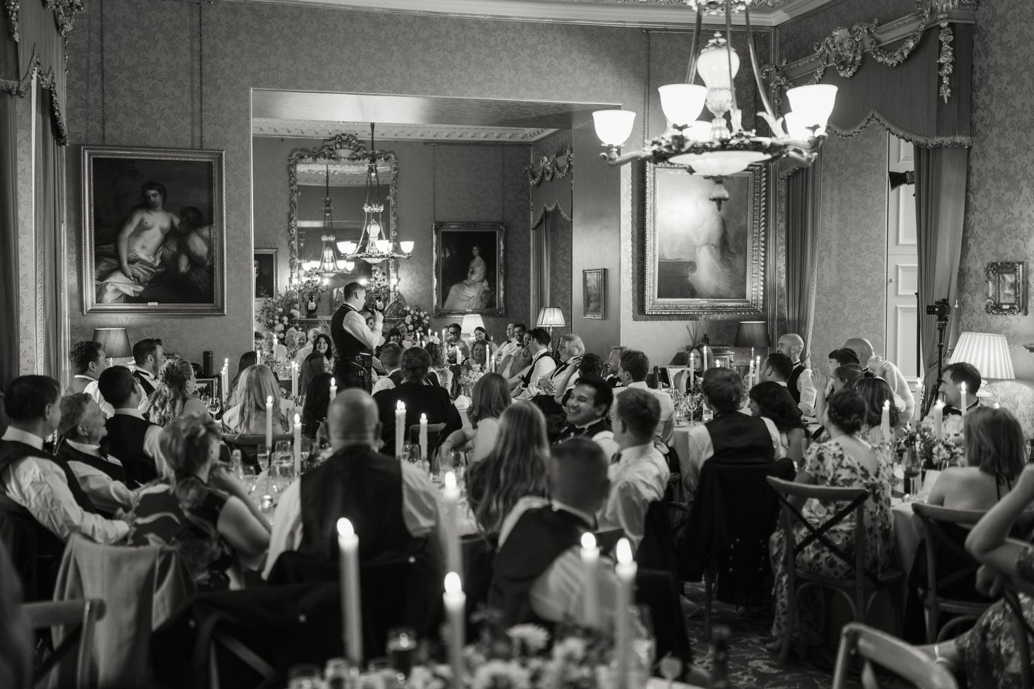 Black and white photo of wedding guests seated at candlelit tables in an elegant dining room with chandeliers and framed paintings.