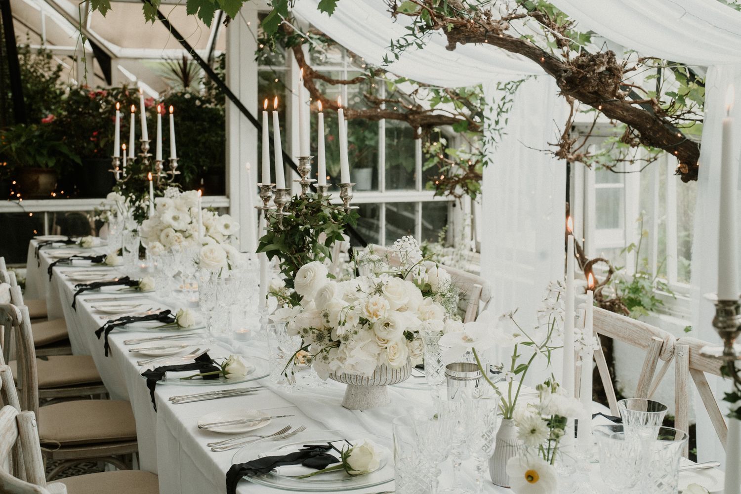 Long wedding reception table set with white linens and floral arrangements in greenhouse setting.