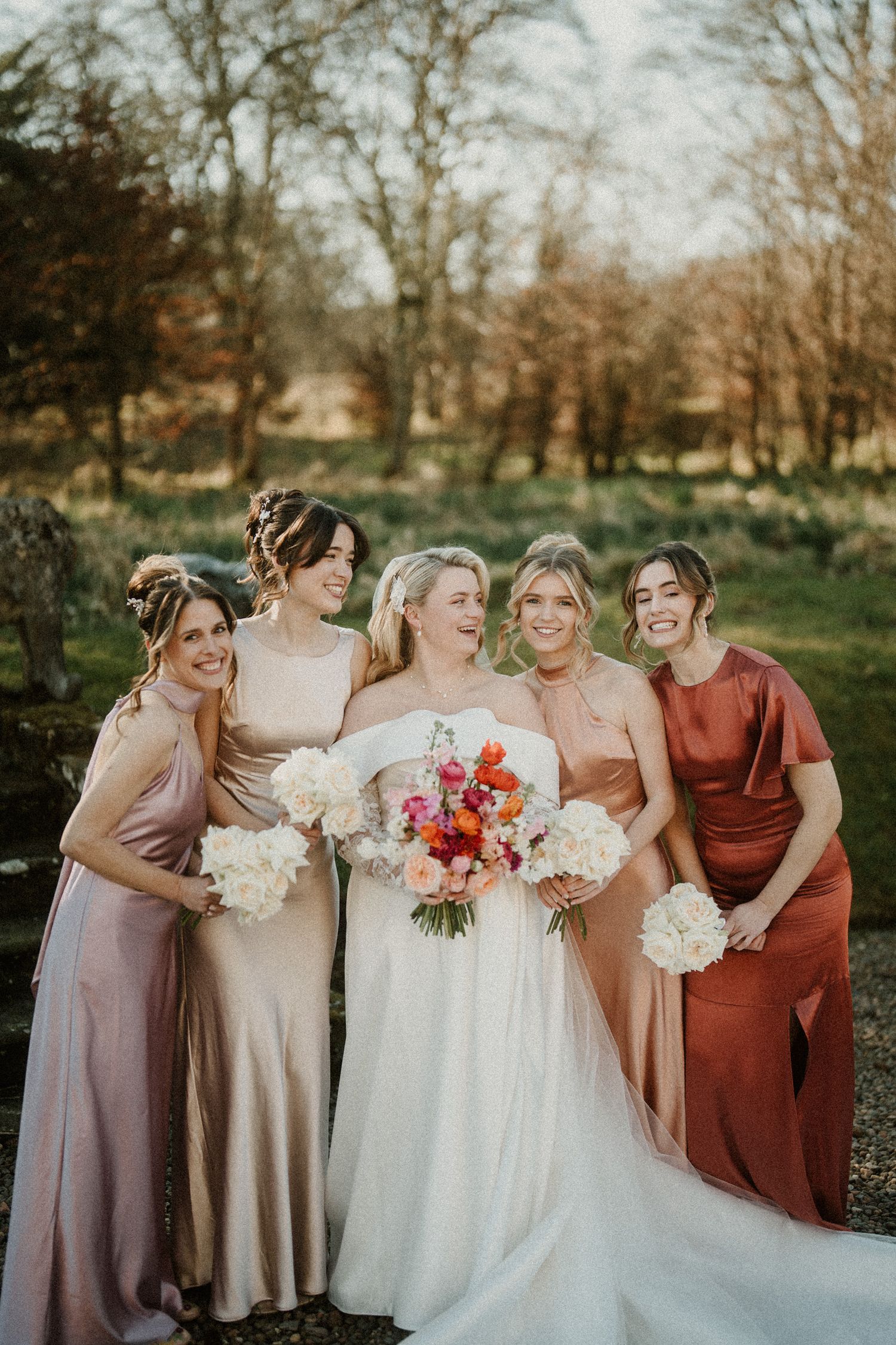 Wedding party holding bouquets poses together in earth-toned dresses during fall celebration.