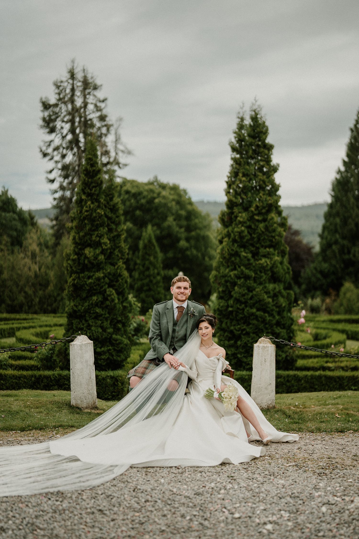Wedding couple seated on stone steps with long flowing veil spread across gravel path.
