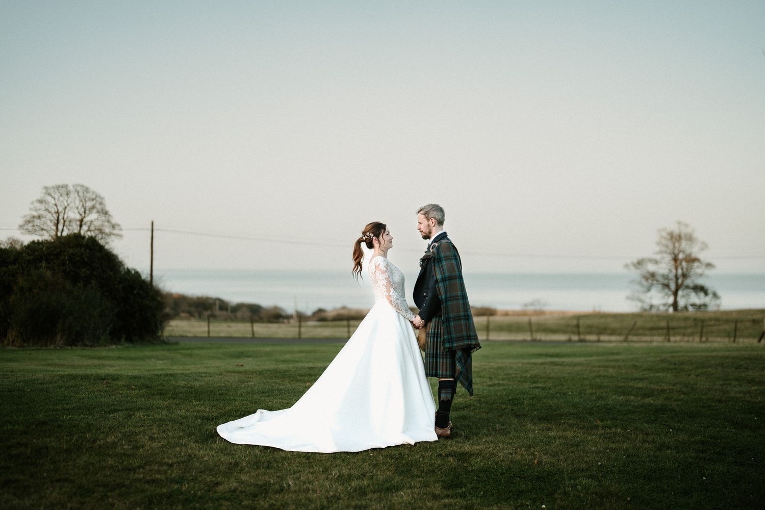 Newlyweds in wedding dress and kilt walking through a scenic field at dusk.