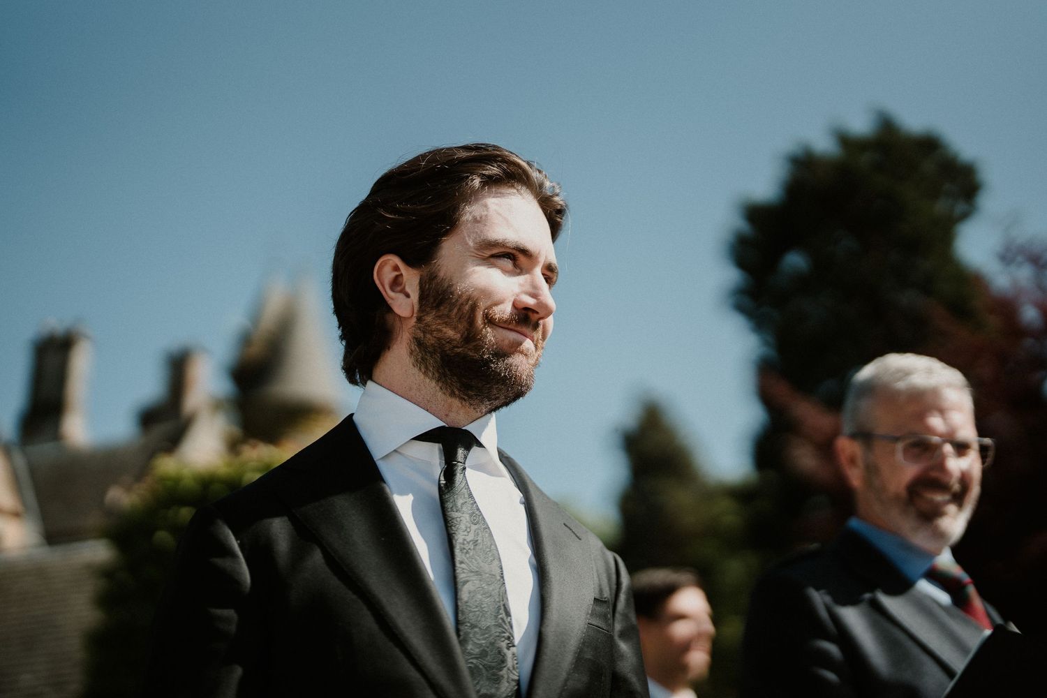 Groomsmen in dark suits standing against bright blue sky at outdoor wedding.