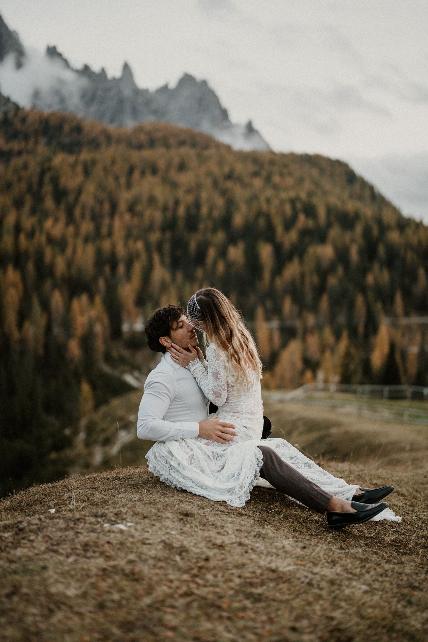 Couple dressed in white embraces on a hillside with golden autumn trees and mountains in the background.