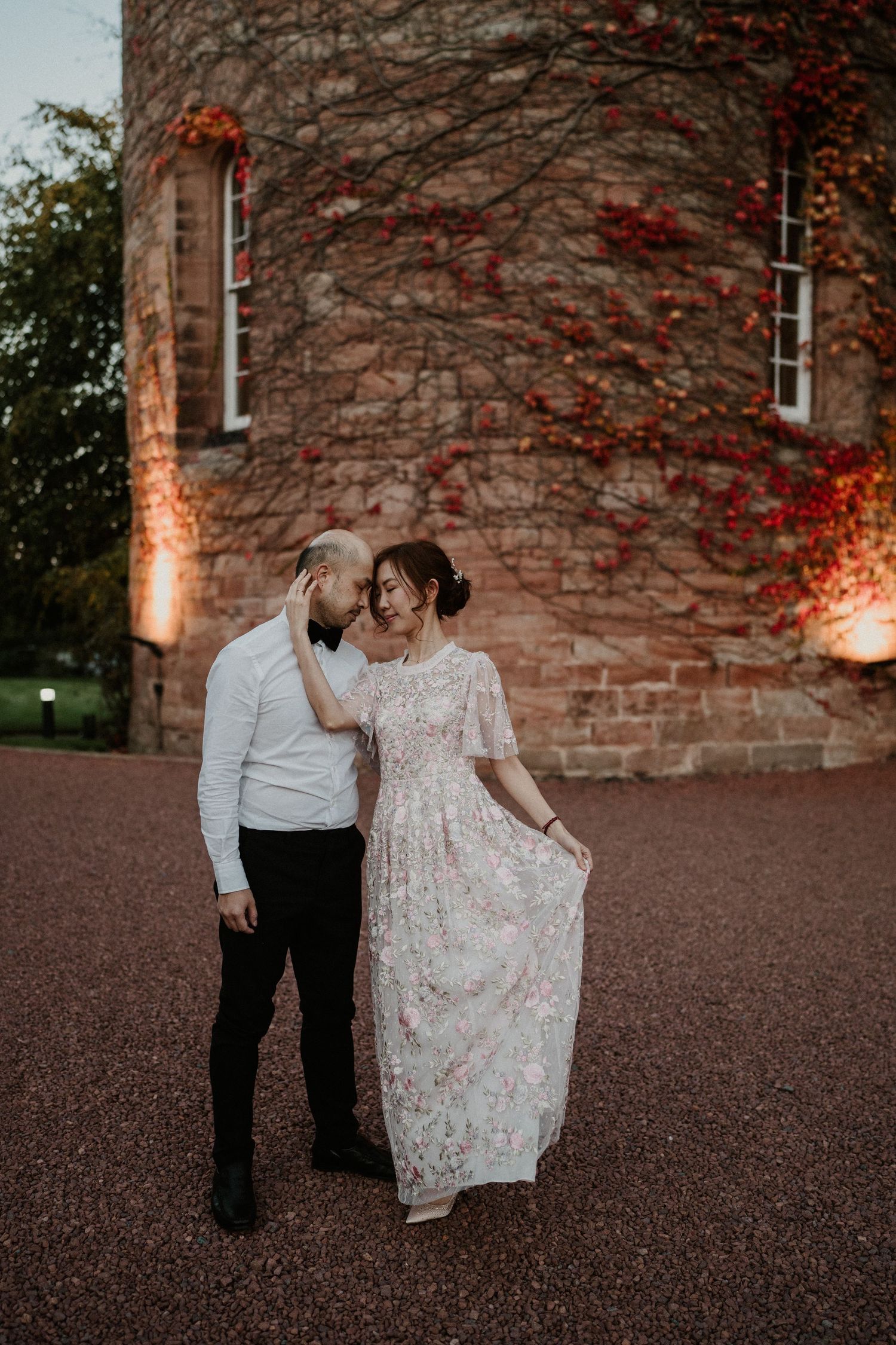 A couple shares an intimate moment against a stone wall with climbing red vines.