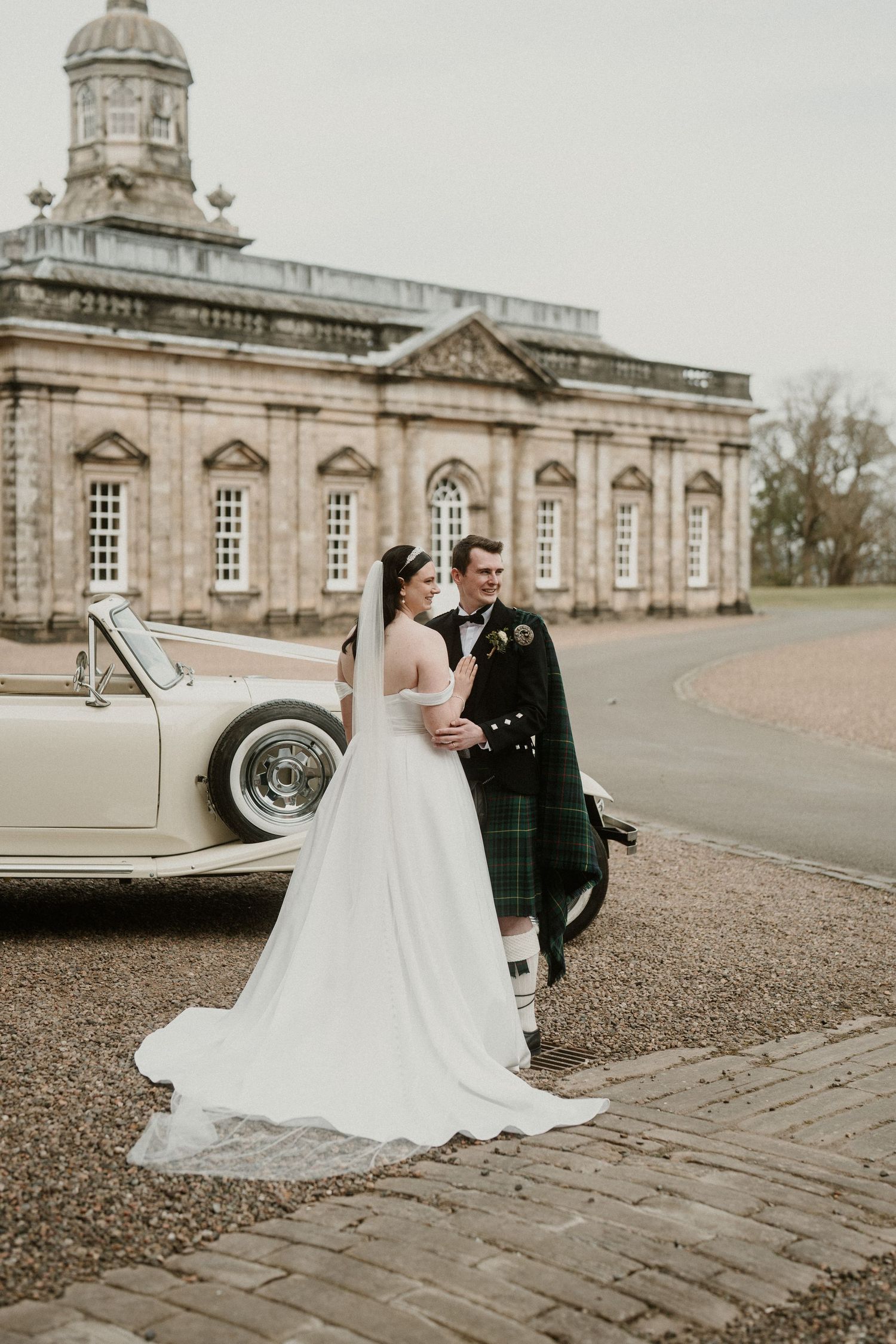Bride in flowing white gown poses with groom in Scottish kilt outside historic building.