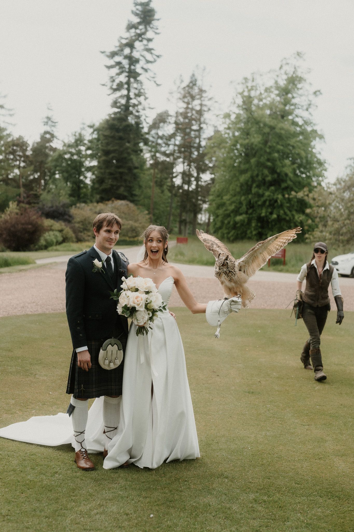 Bride and groom posing outdoors with an owl landing on bride's hand, groom in tartan kilt, holding white bouquet.