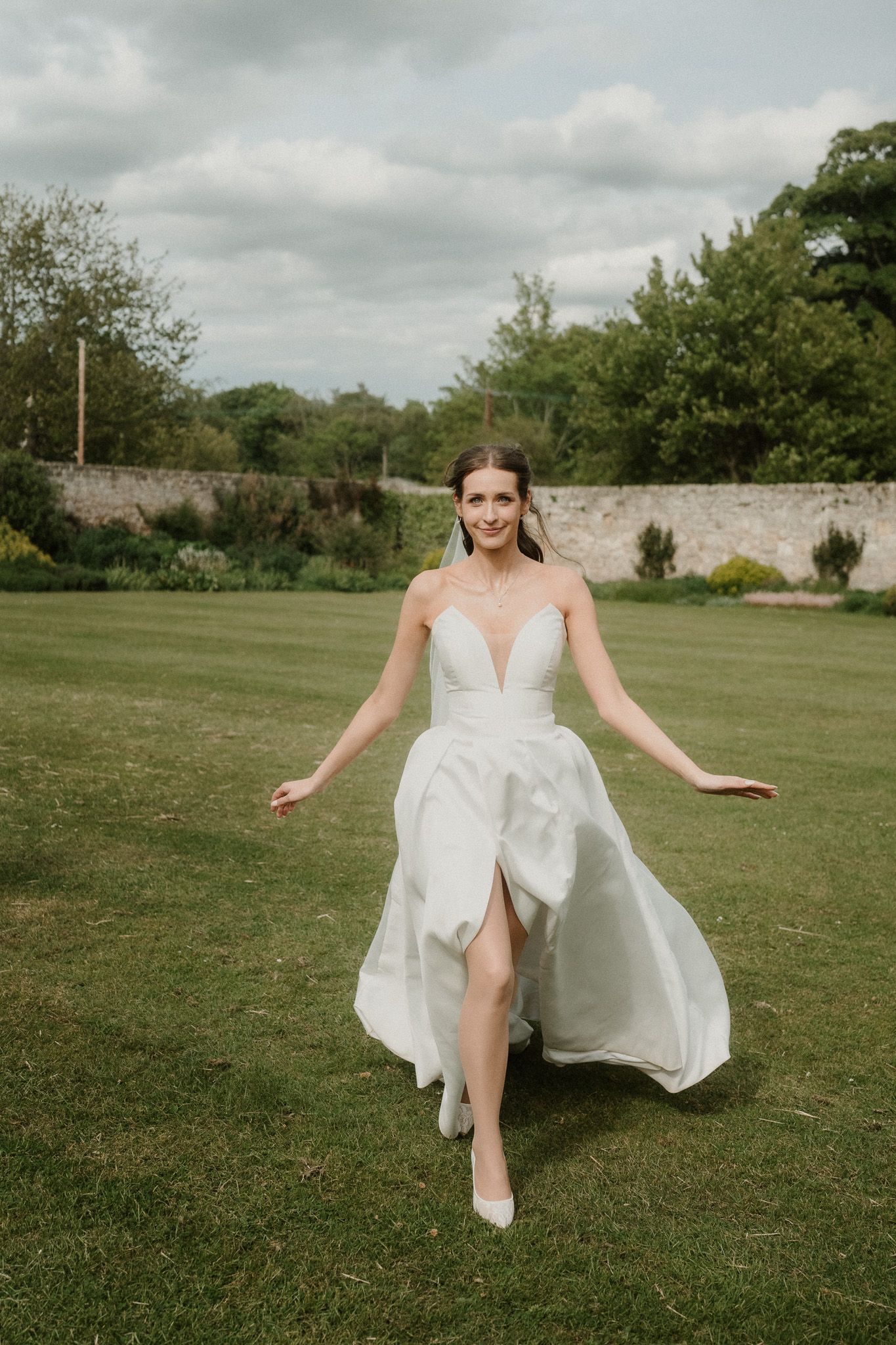 Bride in strapless white wedding gown with deep V-neckline and flowing skirt walks across lush green garden lawn.