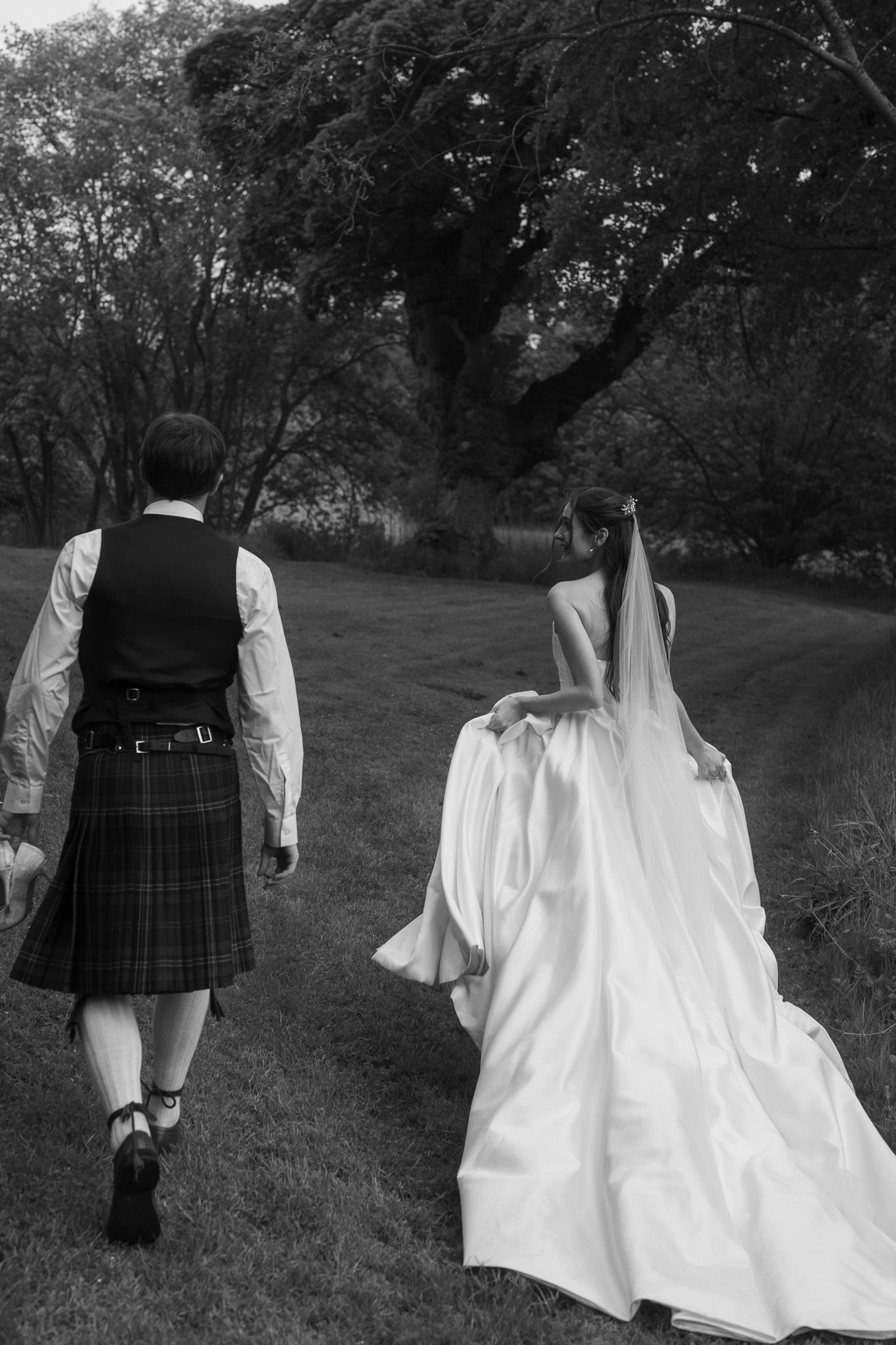 Black and white photo of bride in white gown and groom in tartan kilt walking through lush garden.
