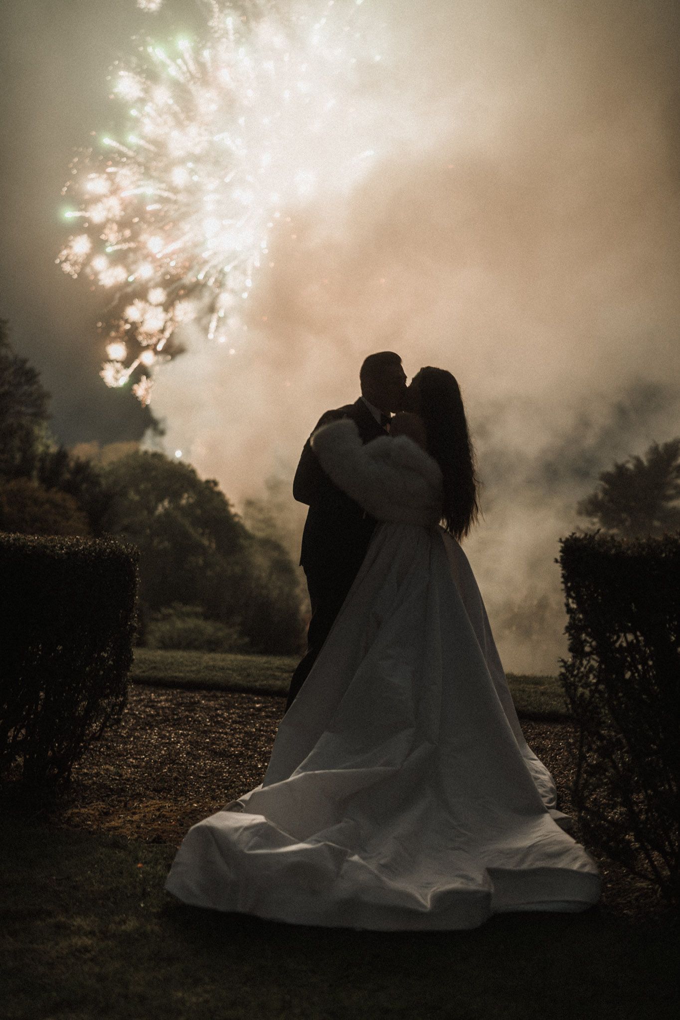 A romantic silhouette of a couple in wedding attire embracing during a misty twilight photoshoot in a garden setting.
