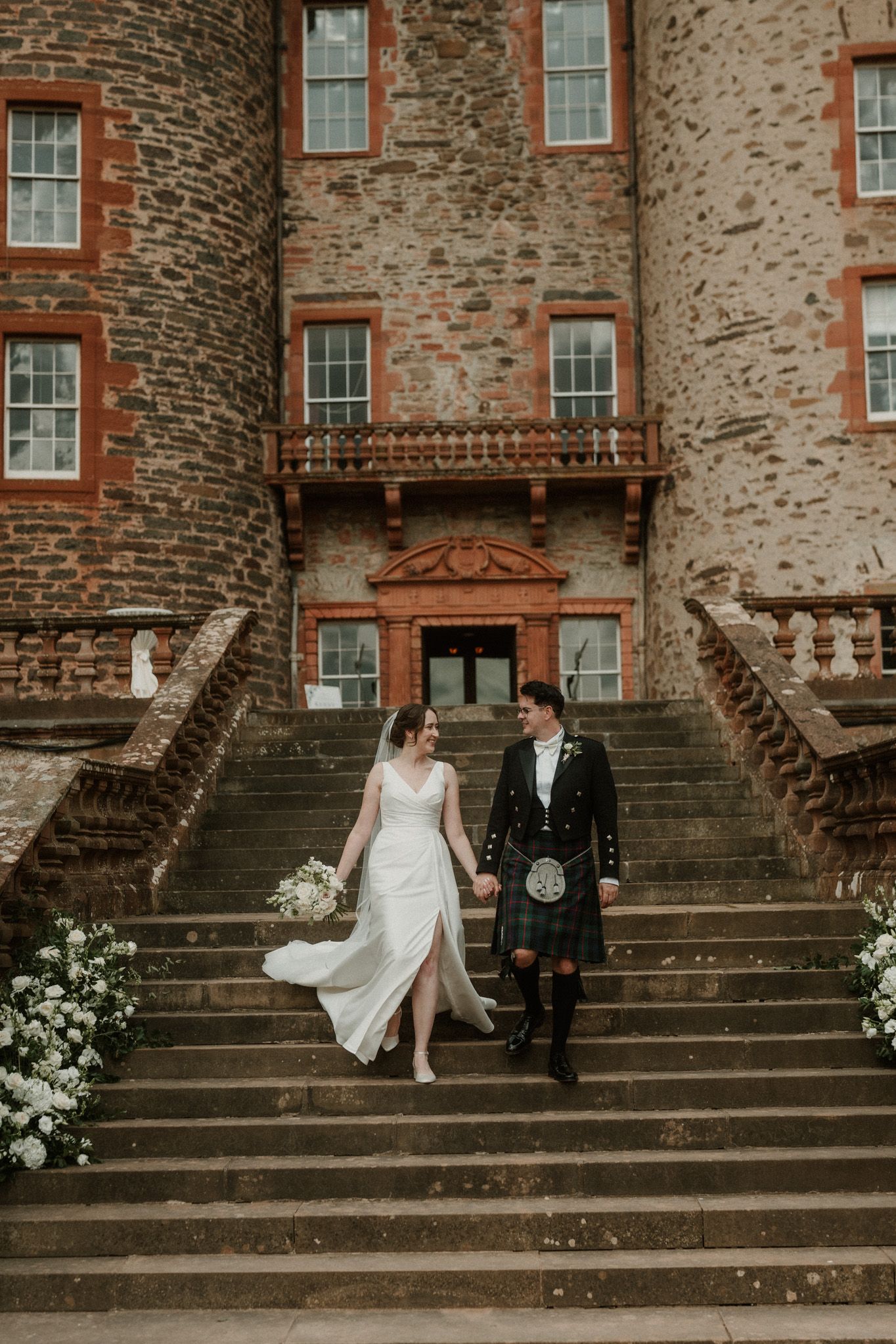 Bride in a white gown and groom in a traditional kilt hold hands while descending stone castle steps at a Scottish wedding.
