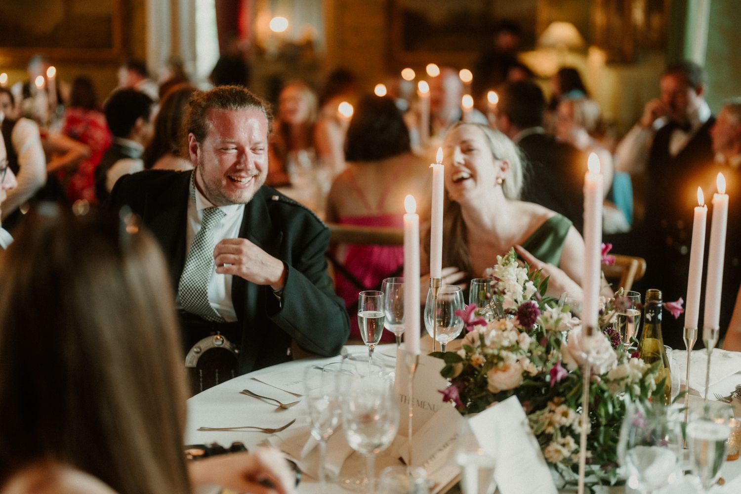 Guests laugh and chat at an elegant candlelit banquet table set with flowers, wine glasses and tall white candles.