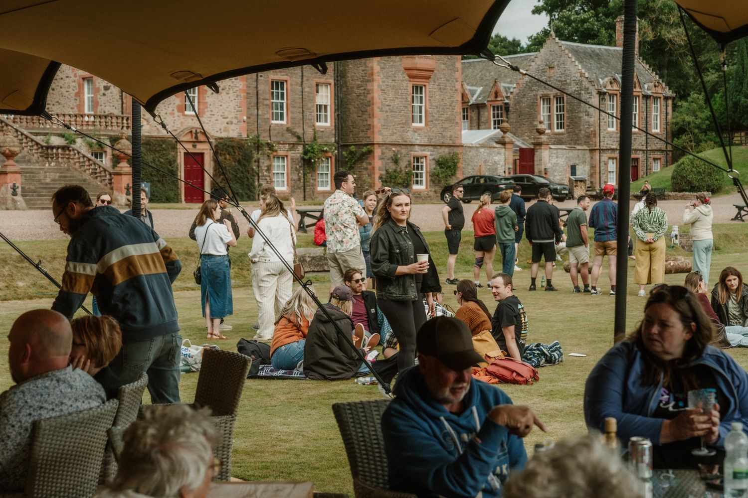 Guests relax and mingle on a sunny lawn beneath a canopy tent, with a historic stone manor house in the background at an outdoor event.