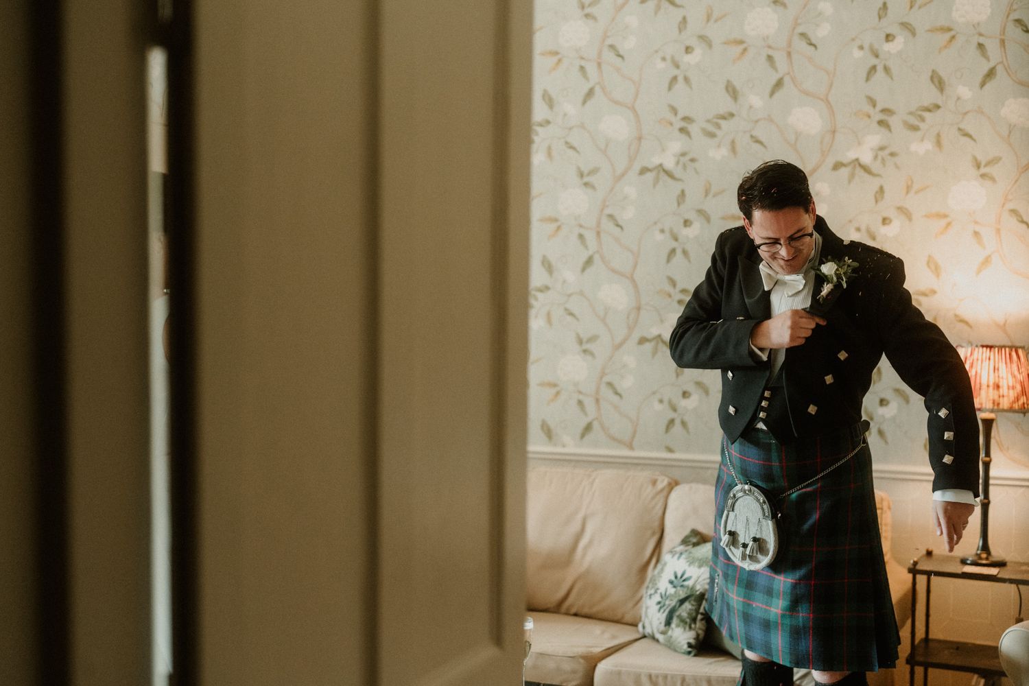 Groom in a black jacket and tartan kilt with sporran adjusts his boutonniere beside a cozy sofa in a softly lit room with floral wallpaper.
