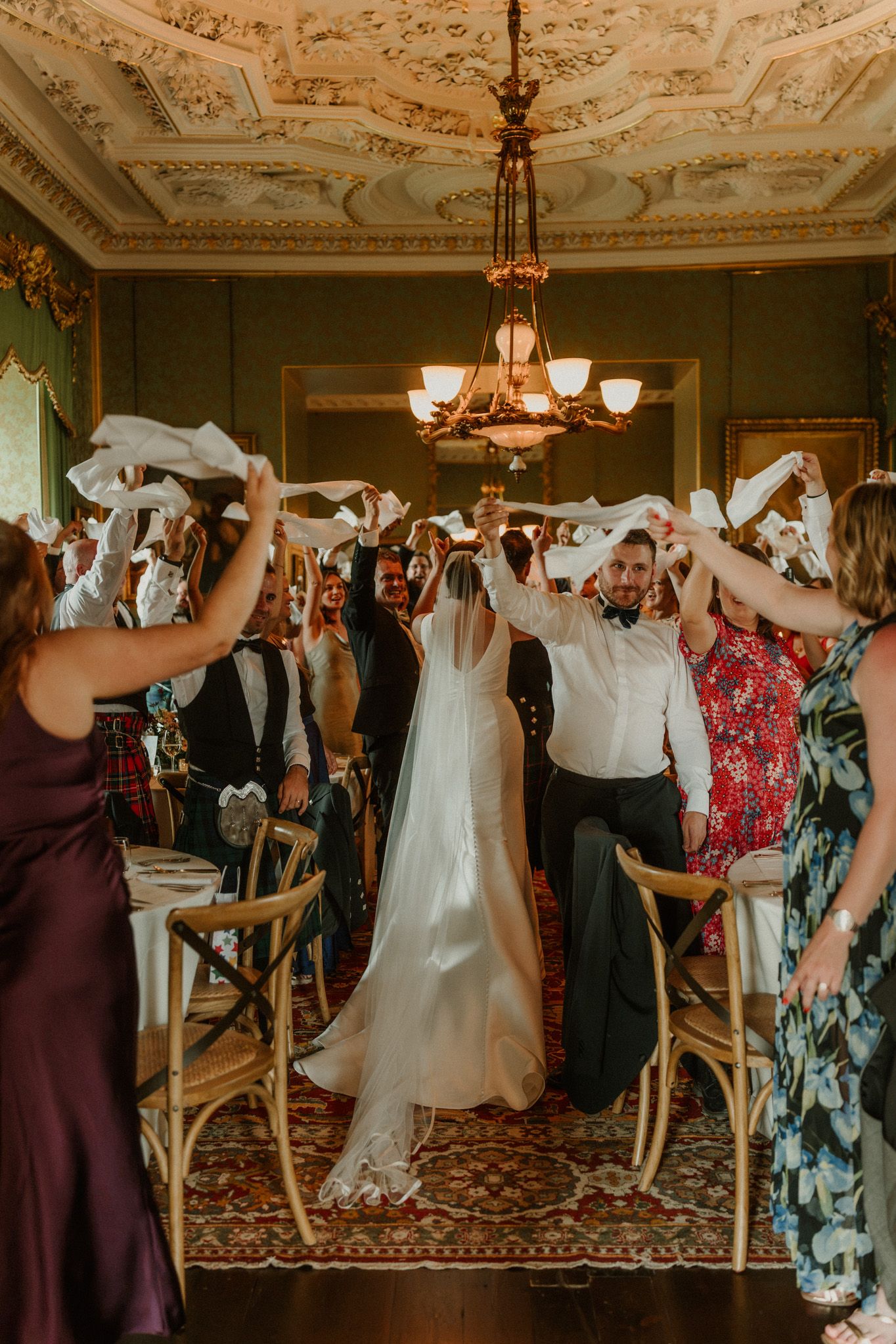 Wedding guests wave white napkins overhead as the bride and groom walk down an elegant ballroom aisle beneath an ornate chandelier.
